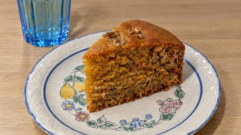 Una porción de bizcocho de zanahoria y avena con nueces por encima, en un plato con cenefa de flores y un vaso de agua al lado, vista en ángulo de 45º