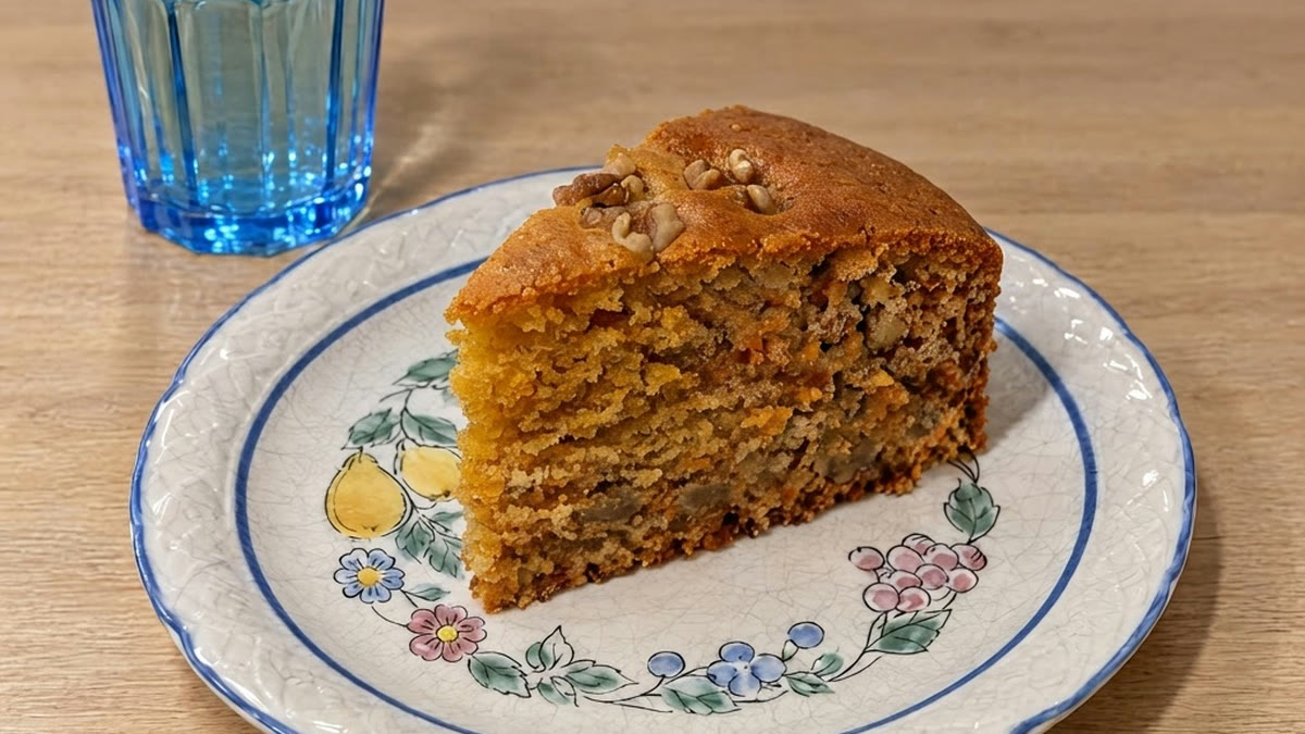 Una porción de bizcocho de zanahoria y avena con nueces por encima, en un plato con cenefa de flores y un vaso de agua al lado, vista en ángulo de 45º