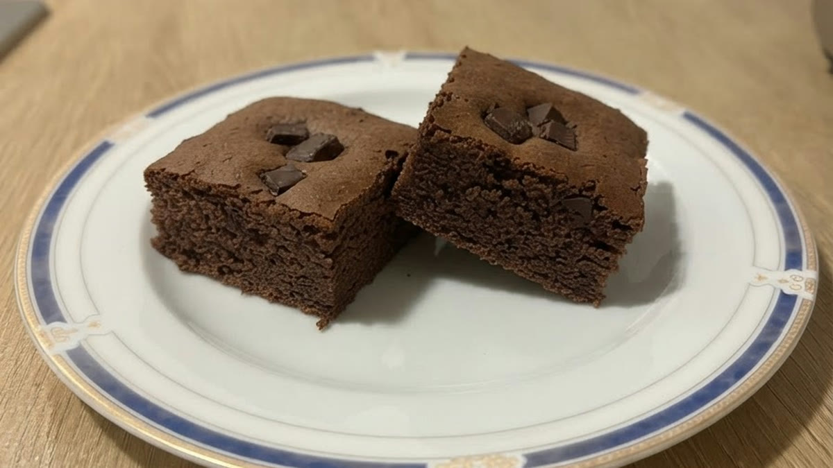 Dos cuadrados de brownie proteico de avena y cacao con trozos de chocolate 85 % por encima, en un plato blanco con borde azul, vistos a 45º