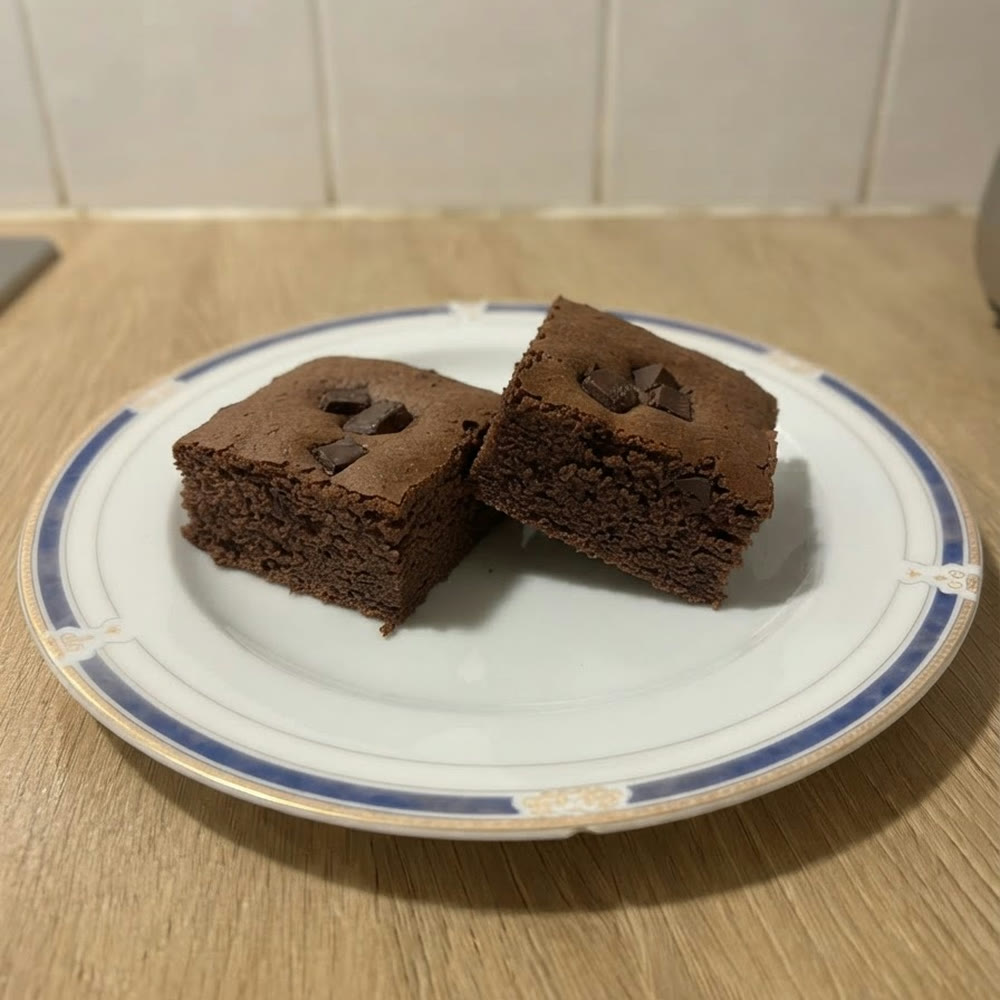 Dos cuadrados de brownie proteico de avena y cacao con trozos de chocolate 85 % por encima, en un plato blanco con borde azul, vistos a 45º