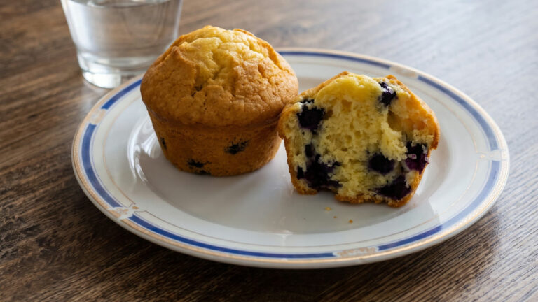 Plato con dos muffins de avena y arándanos con proteína, uno entero y otro partido con los arándanos a la vista, y un vaso de agua detrás, sobre una mesa de madera