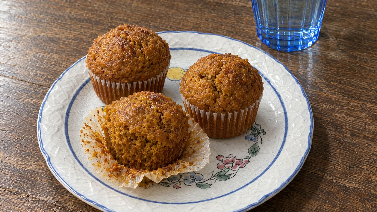Tres muffins de calabaza y avena en un plato blanco con flores pintadas, junto a un vaso azul con agua, sobre una mesa de madera