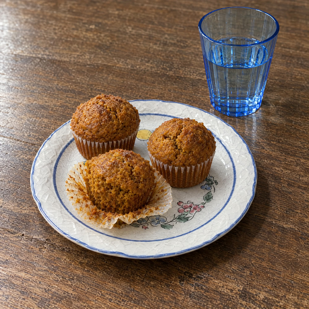 Tres muffins de calabaza y avena en un plato blanco con flores pintadas, junto a un vaso azul con agua, sobre una mesa de madera