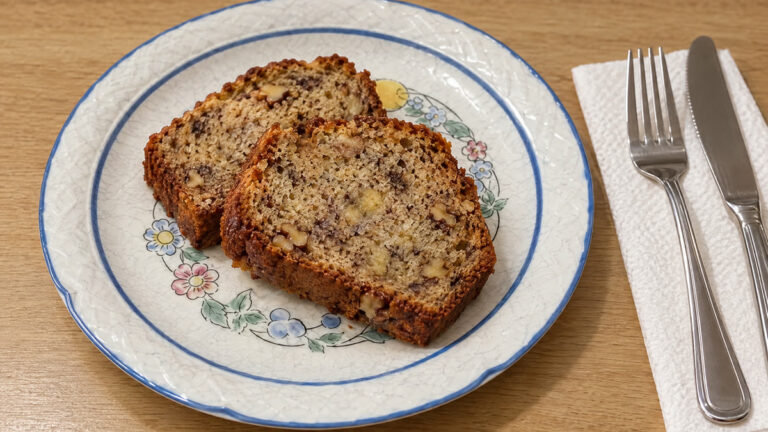 Dos rebanadas de pan de plátano fitness con avena y nueces en un plato blanco de borde azul, con tenedor y cuchillo al lado, vistas en ángulo de 45 grados